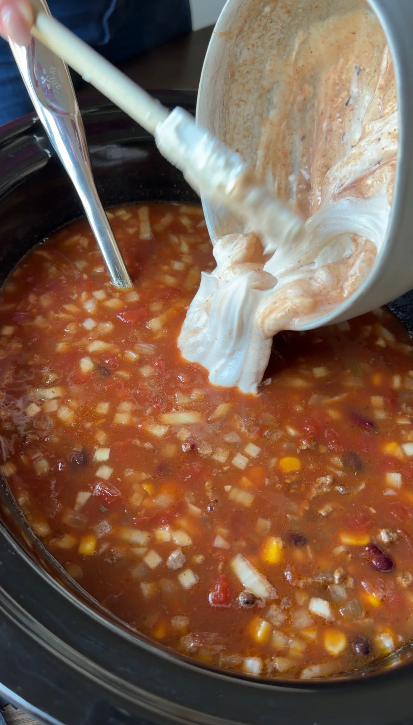 A hand uses a spatula to scrape a creamy white mixture from a bowl into a slow cooker filled with chunky vegetable and bean soup.