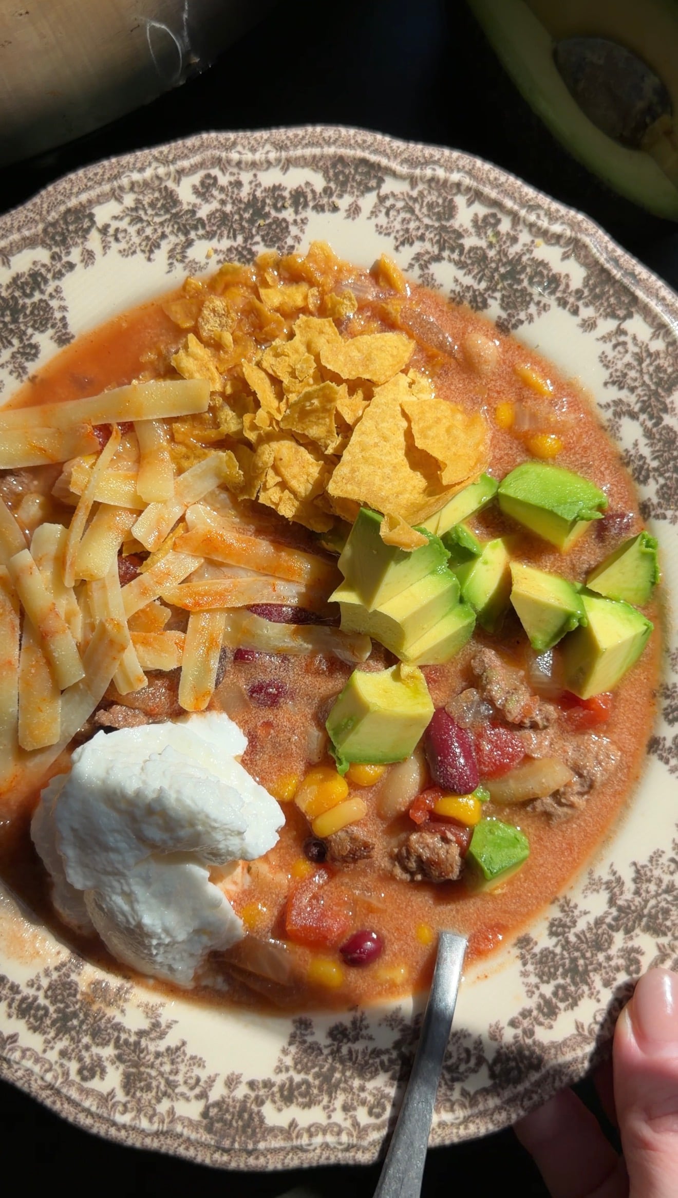 A bowl of chili topped with shredded cheese, tortilla chips, diced avocado, and a dollop of sour cream.