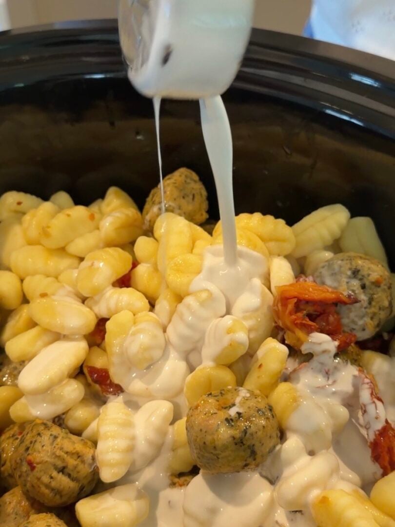 Cream being poured from a measuring cup over a mixture of gnocchi, meatballs, and sun-dried tomatoes in a slow cooker.