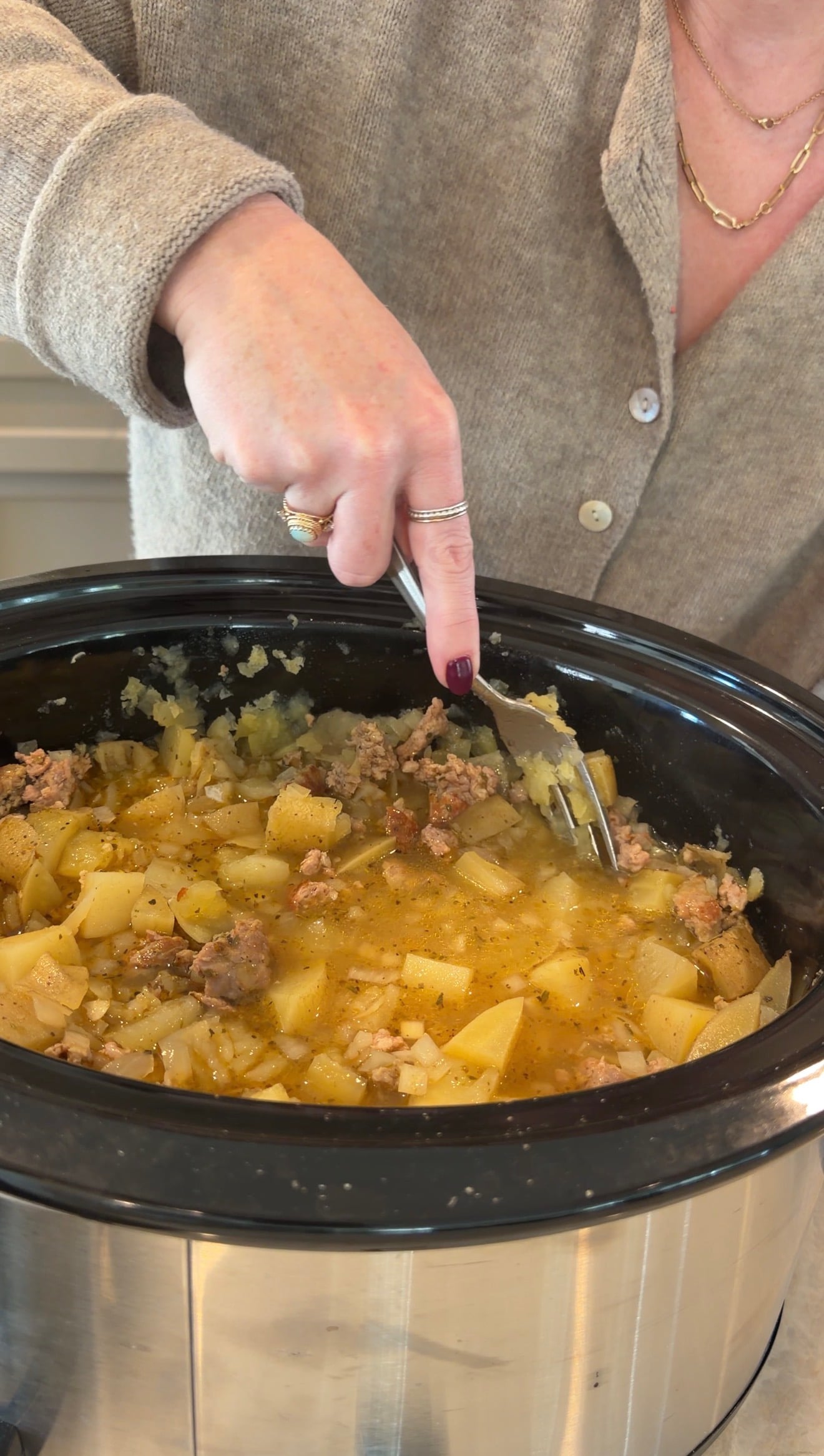 A hand uses a fork to stir a mixture of chopped potatoes and ground meat in a slow cooker.