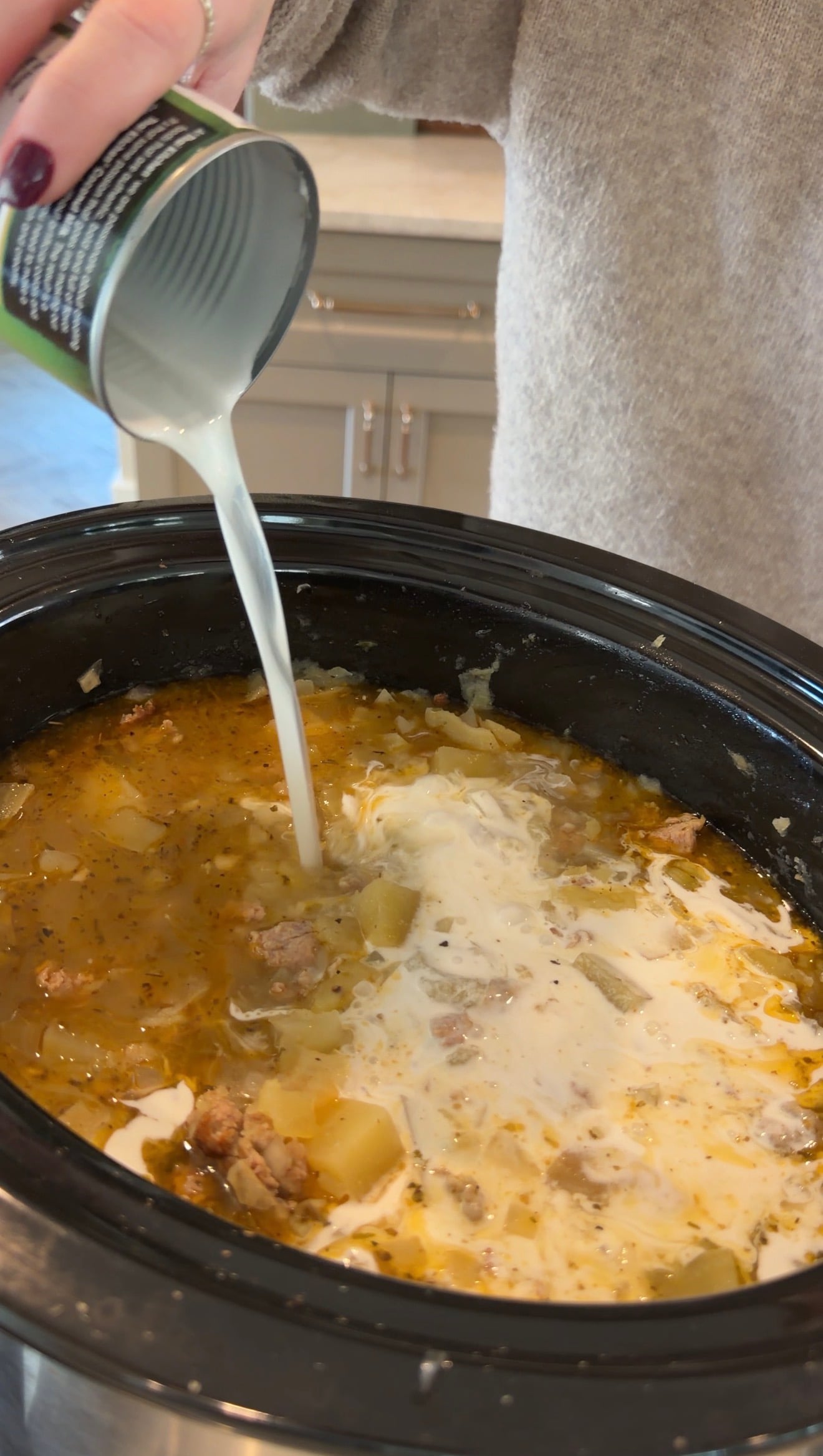 A person pours a can of cream into a slow cooker filled with potato and sausage soup.