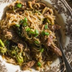 A bowl of beef and broccoli stir-fry with noodles, topped with sesame seeds and green onions.