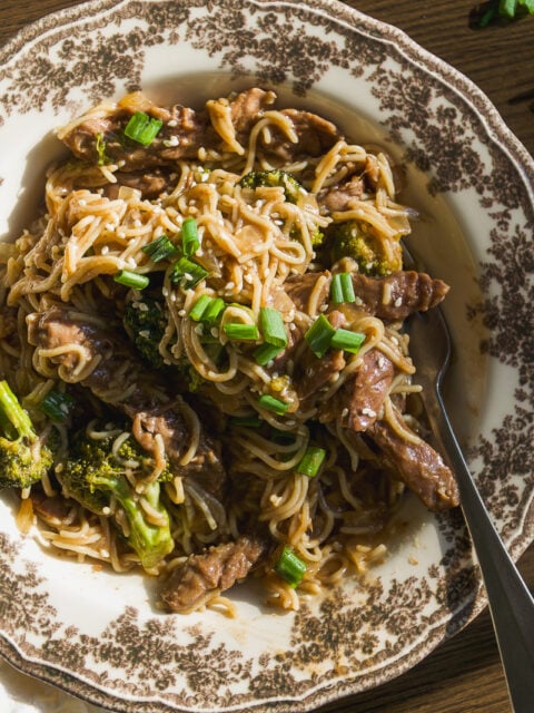 A bowl of beef and broccoli stir-fry with noodles, topped with sesame seeds and chopped green onions.