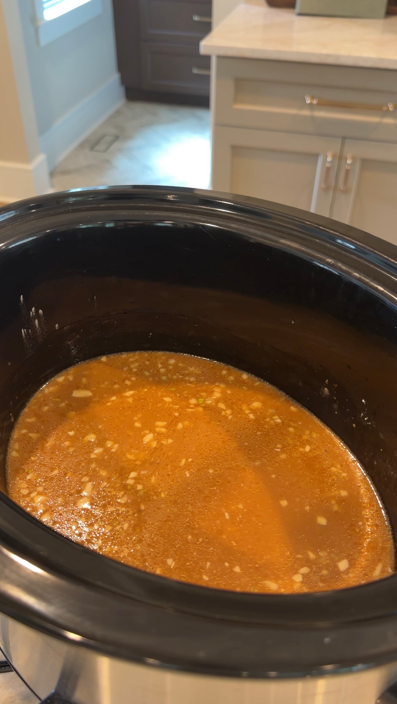 A slow cooker filled with a reddish-brown broth containing small bits of garlic or onion.