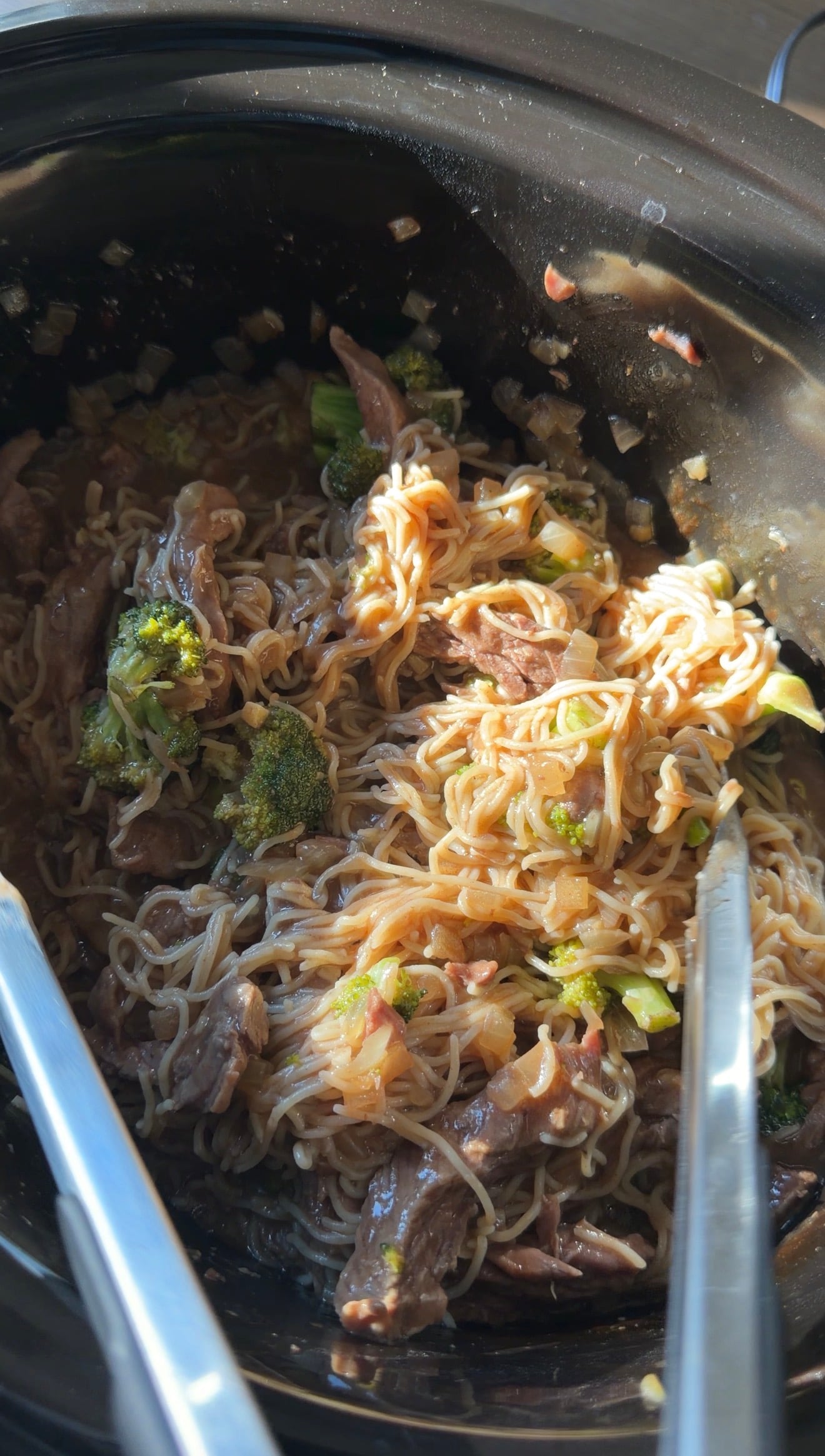 A slow cooker filled with a mixture of beef, broccoli, and thin noodles in a brown sauce, with metal tongs resting inside.