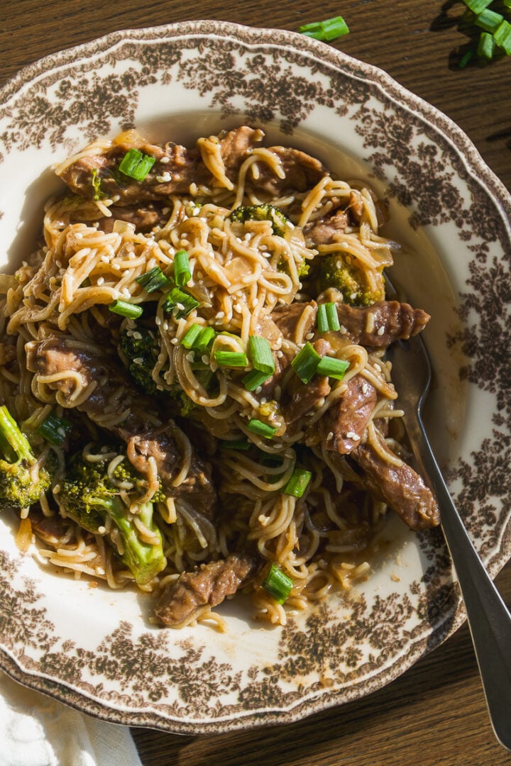 A bowl of beef and broccoli stir-fry with noodles, topped with sesame seeds and chopped green onions.