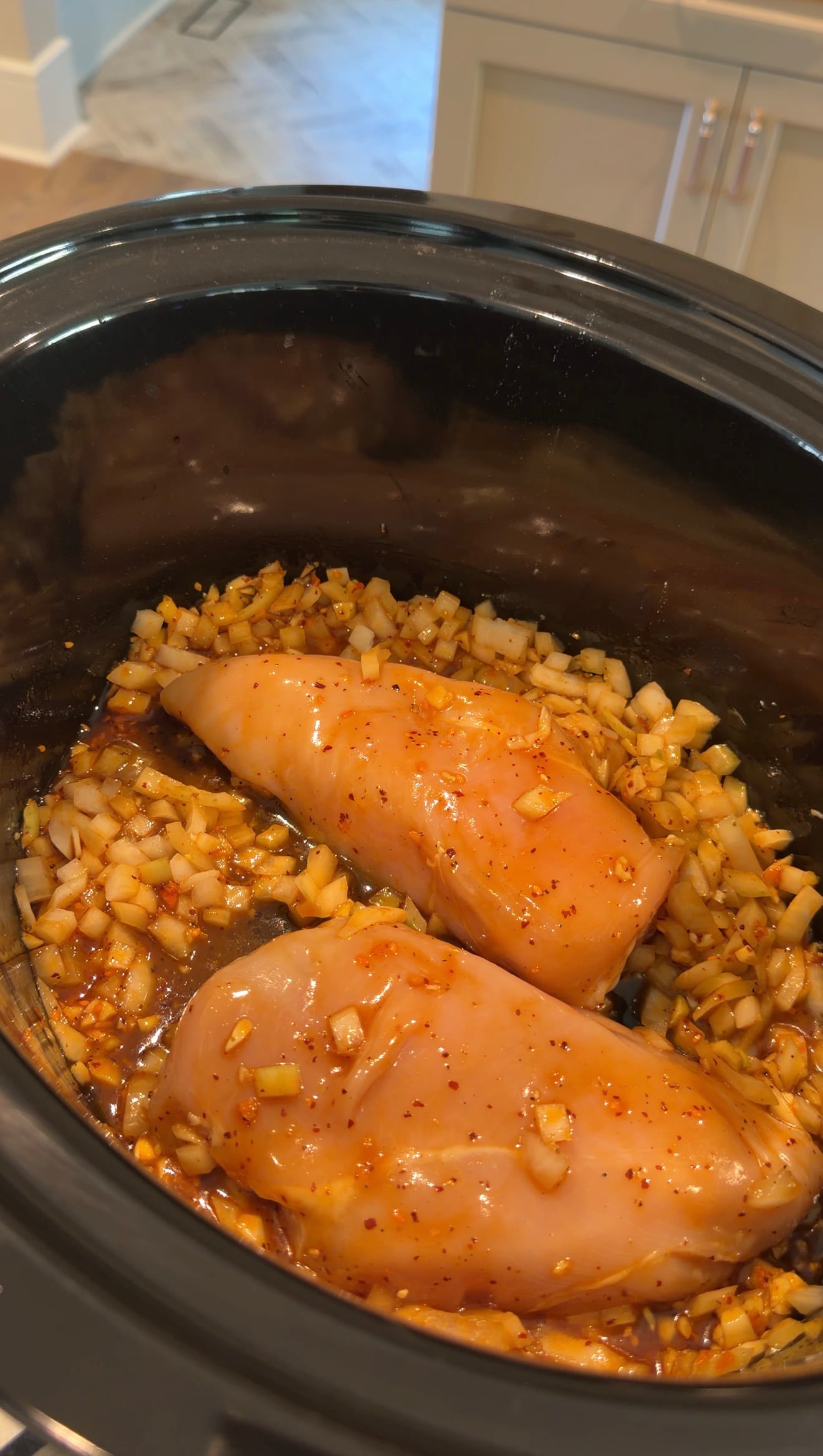 Two raw chicken breasts cooking in a slow cooker with diced onions and a red sauce.