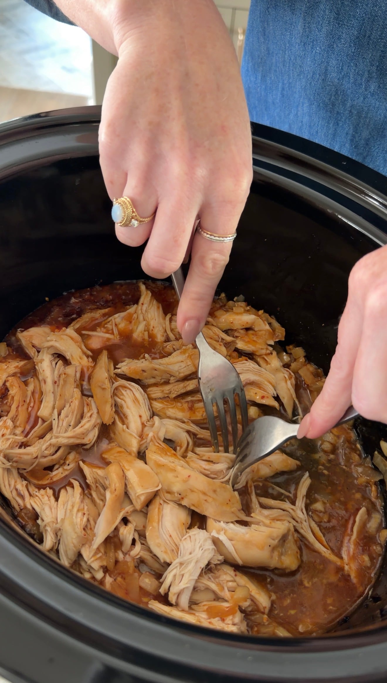 A person uses two forks to shred cooked chicken in a slow cooker.