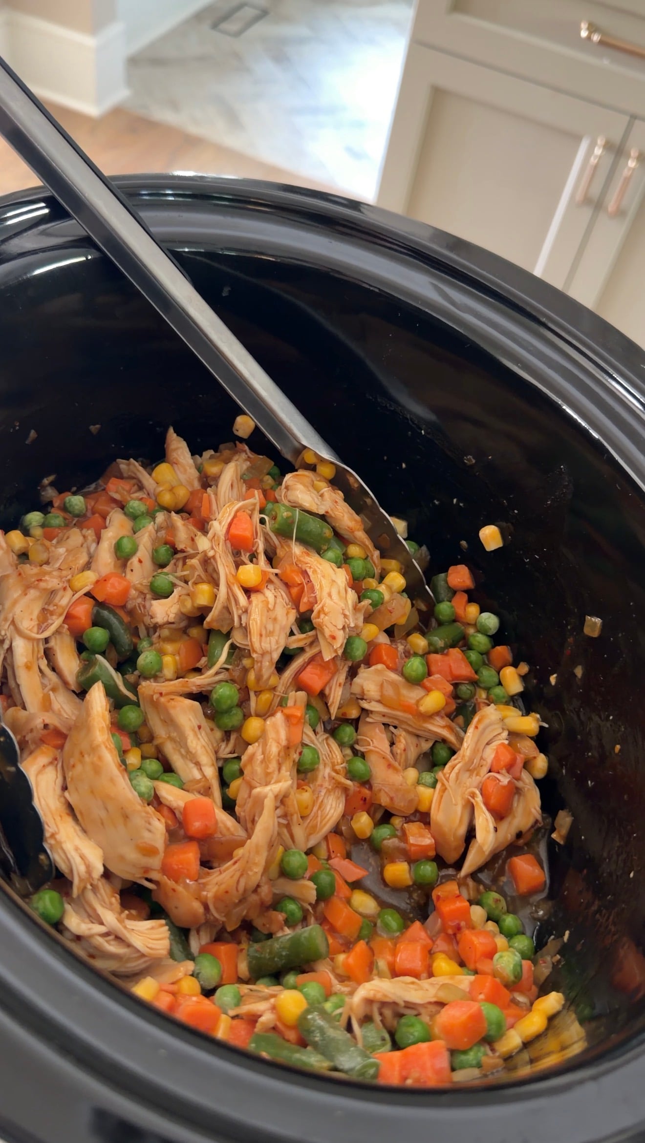 Shredded chicken mixed with peas, corn, and carrots in a slow cooker with a serving spoon.