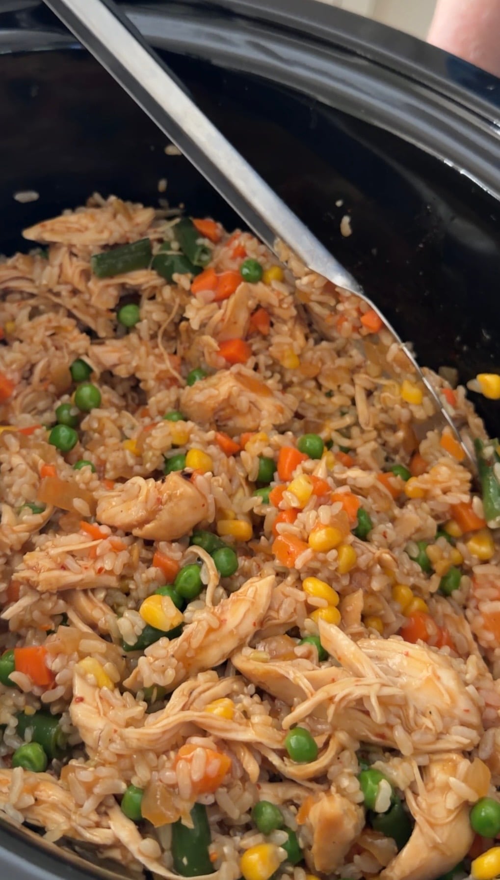 A serving spoon in a slow cooker filled with chicken and vegetable fried rice containing corn, peas, carrots, and green beans.