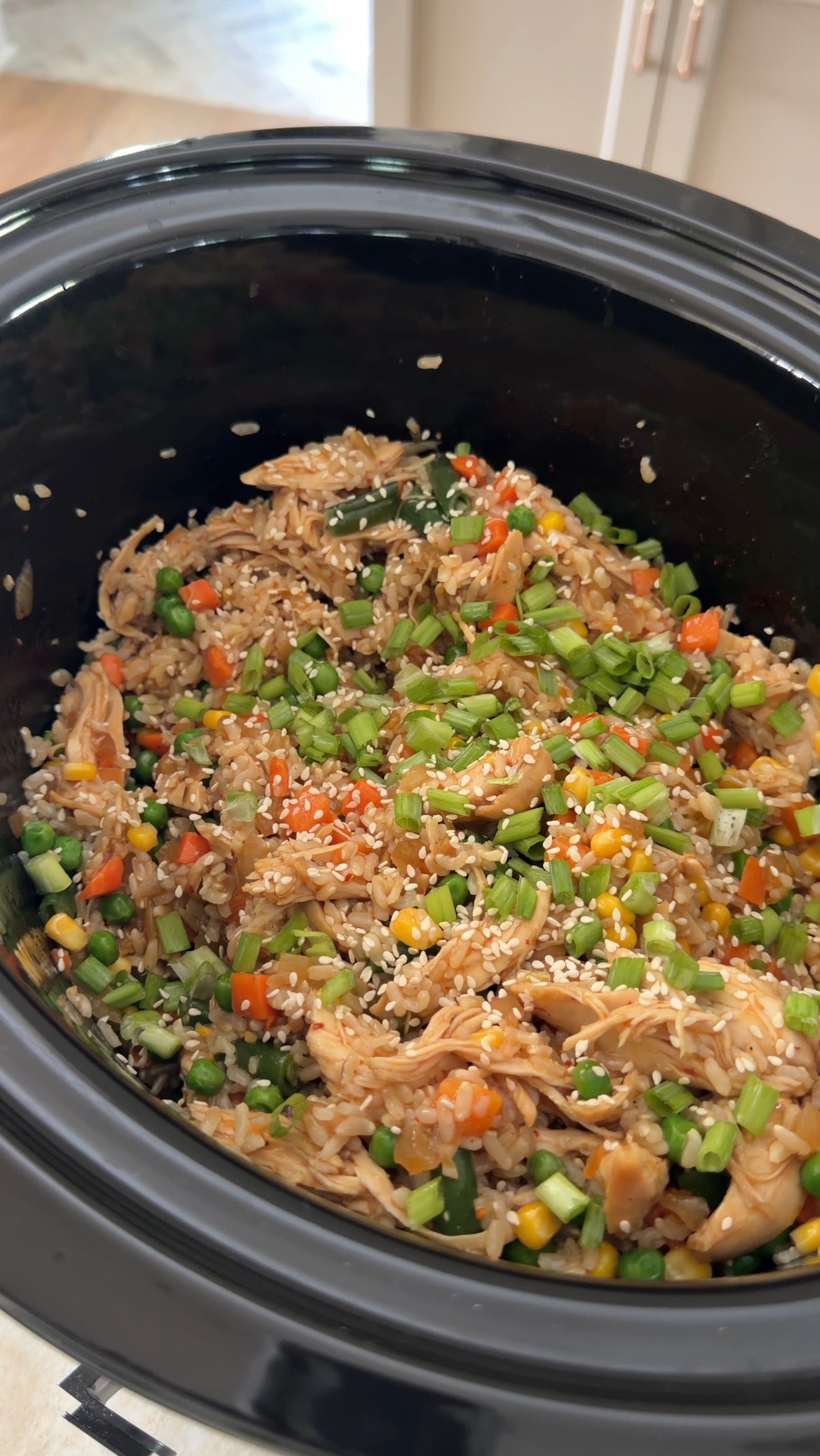 A slow cooker filled with chicken, rice, corn, peas, and carrots, garnished with sesame seeds and chopped green onions.