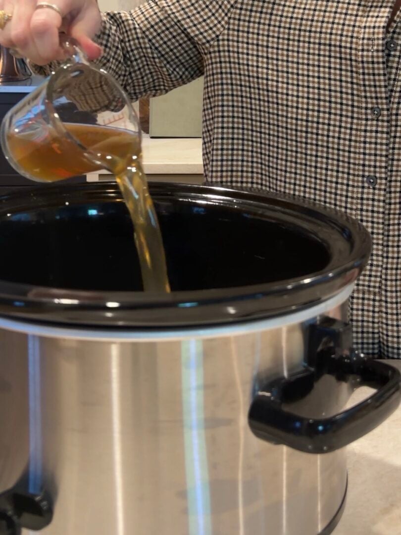 A person pouring broth from a glass measuring cup into a slow cooker.