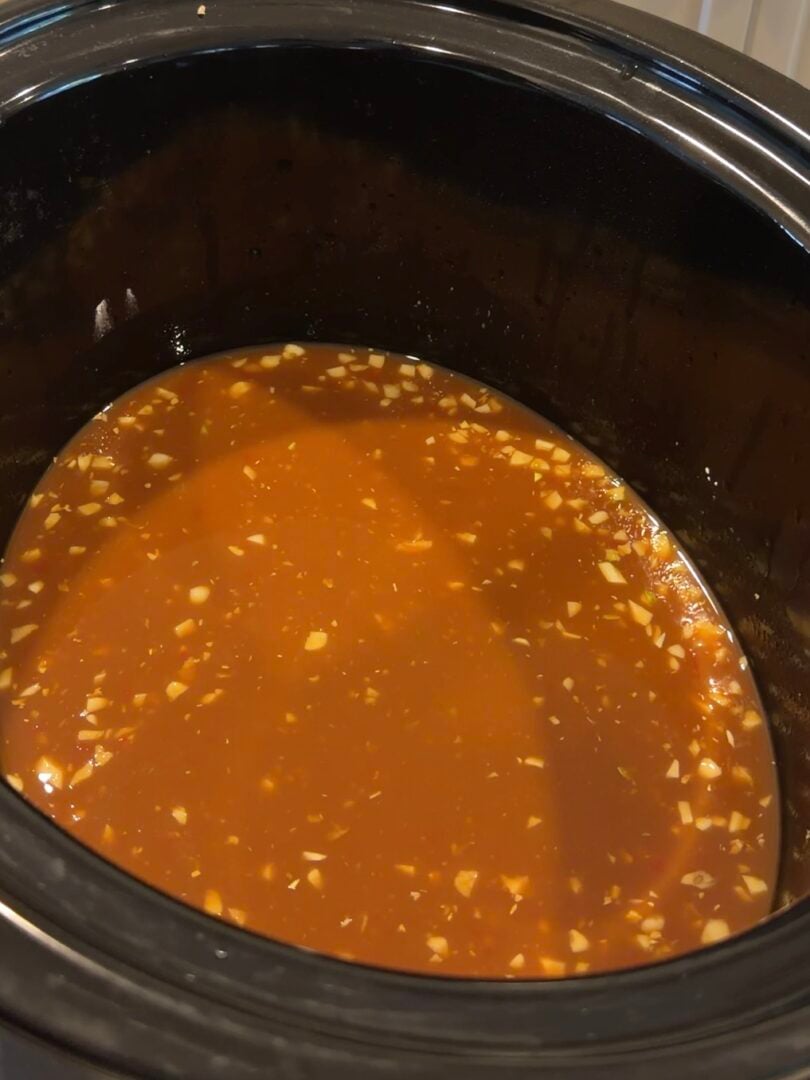 A brown, broth-based soup with visible pieces of minced garlic cooking in a black slow cooker.