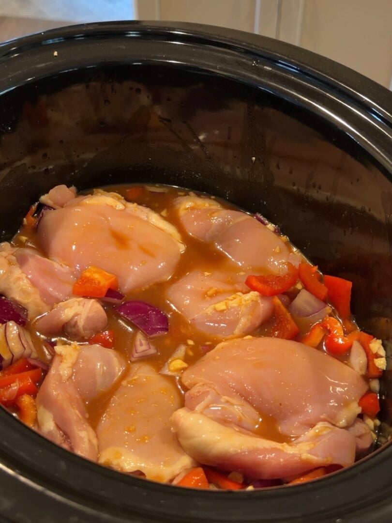 Raw chicken breasts with chopped red bell peppers and red onions in a slow cooker filled with sauce.