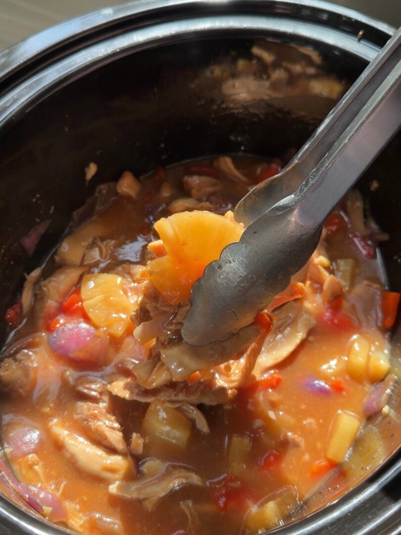 Metal tongs holding a piece of pineapple and shredded chicken from a slow cooker filled with a stew of chicken, pineapple, and red onion.